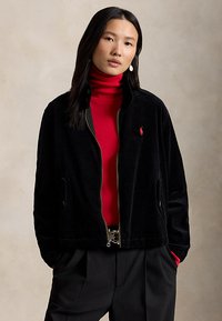 Femeie cu păr lung și închis la culoare, purtând o jachetă neagră de catifea cu dungi peste un pulover roșu cu guler înalt și pantaloni negri, cu mâinile în buzunare.