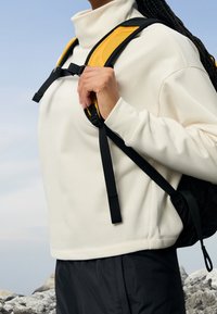 Valkoinen fleece-huppari, jossa korkea kaulus ja löysä istuvuus, yhdistetty mustaan reppuun, jossa on keltaiset pehmustetut olkahihnat ja säädettävä musta nailonhihna.