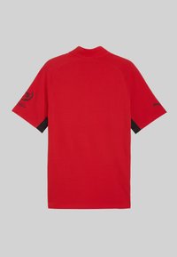 Camiseta roja de manga corta hecha de algodón; presenta un cuello negro y detalles negros en las mangas. Texto y logo estampados en una manga.