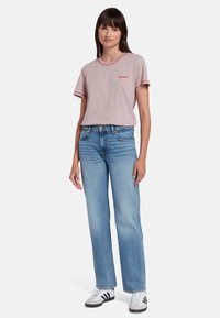 T-shirt beige a maniche corte con bordi e logo rossi, abbinato a jeans straight-leg blu chiaro e sneaker nere e bianche.