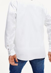 Camisa blanca de manga larga con textura suave, puños abotonados y dobladillo curvado, combinada con jeans de mezclilla azules y un gesto de mano visible.