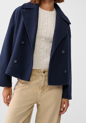 Vrouw die een donkerblauwe double-breasted jas draagt, een crèmekleurige kabelgebreide trui en beige broek met hoge taille, staand met ontspannen handen.