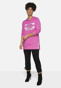 Sudadera con capucha rosa con texto gráfico en la parte frontal, combinada con pantalones negros cortos que presentan detalles de cordones en el dobladillo y botas de tacón alto.