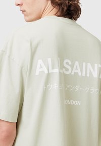 Šviesiai žalia medvilninė marškinėliai su baltu tekstu nugarinėje dalyje, kuriuose parašyta "ALLSAINTS" ir japonų simboliai, apvaliu kaklu ir trumpomis rankovėmis.