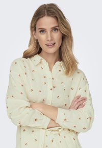 Chemise boutonée jaune clair faite d'un tissu texturé, ornée de petits motifs floraux rouges et verts, à manches longues et avec col.