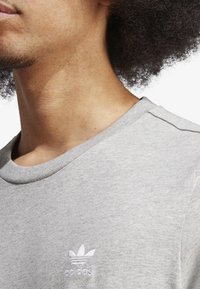 Šedé bavlněné tričko s kulatým výstřihem, na levé straně hrudi s malým bílým vyšívaným logem a jemně strukturovanou texturou.