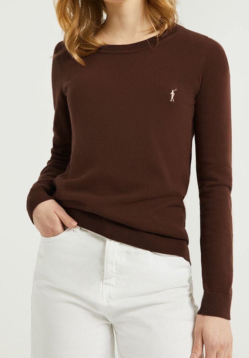 Femme portant un pull brun moulant à manches longues avec un petit logo de golfeur blanc brodé, associé à un pantalon blanc, main dans la poche.