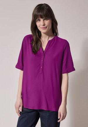 Chemise violette à manches courtes avec une coupe légèrement ample, dotée d'un col en V et de trois boutons sur le devant, fabriquée en tissu lisse.