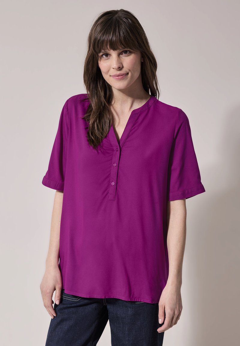 Camicia viola a maniche corte con una vestibilità leggermente ampia, dotata di colletto a scollatura e tre bottoni sul davanti, realizzata in tessuto liscio.