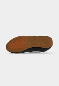 Basket noire avec une semelle en caoutchouc marron montrant un motif de bande de roulement en zigzag, vue par le dessous sur fond blanc.