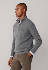 Maglione grigio con zip a metà colletto, lavorazione a maglia testurizzata, polsini e orlo a costine. Indossato sopra una camicia a righe blu chiaro e bianco.