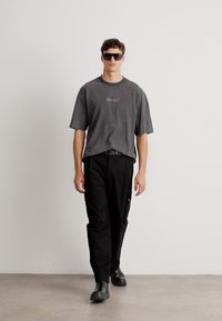 Graues, übergroßes T-Shirt mit "HUGO"-Logo, kombiniert mit schwarzen Cargo-Hosen. Model trägt schwarze Sonnenbrille und schwarze Stiefel und steht vor einem hellen Hintergrund.