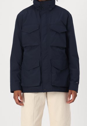 Man wearing navy blue jacket with four large flap pockets and beige pants, standing against plain white background.