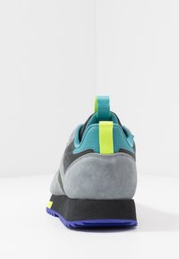 Šedý semišový teniska s černou podrážkou, zelenými a modrozelenými akcenty, poutkem a texturovaným vzorem. Má nízký střih.