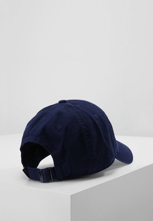 Casquette de baseball bleu marine avec une sangle ajustable et une boucle métallique posée sur une surface blanche sur un fond gris clair.