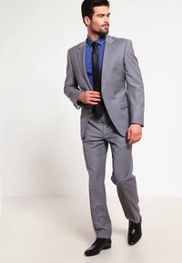 Pilkas kostiumas, pritaikytas kūno linijoms, derinamas su mėlyna marška ir juoda kaklaraiščiu. Juodi oficialūs batai užbaigia įvaizdį. Lygus audinys be matomų raštų.