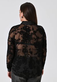 Black sheer blouse with floral embroidery, long sleeves, and a collar. Features a smooth, textured surface and a slightly loose fit.