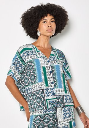 Frau mit Afro-Frisur, trägt große silberne Ohrringe, ein gemustertes blau-grünes Hemd mit Knöpfen, passende Hosen und ein silbernes Armreif.