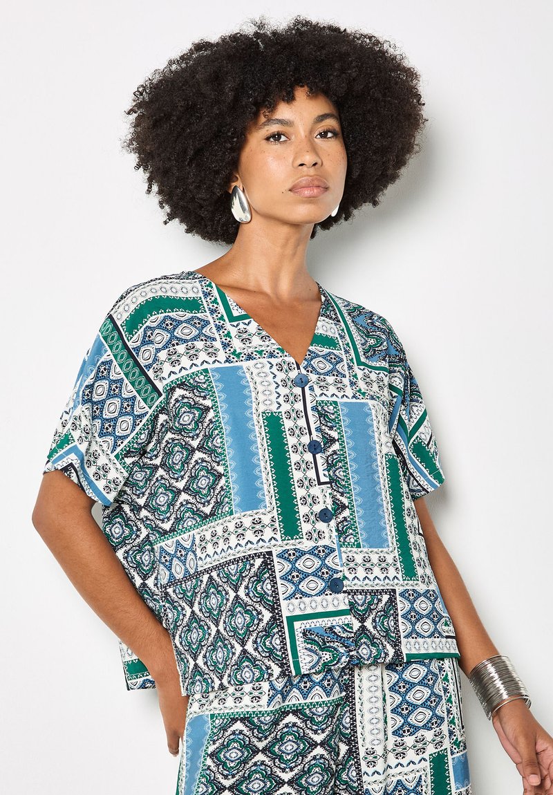 Femme avec une coiffure afro portant de grandes boucles d'oreilles argentées, un haut boutonné à motifs bleus et verts, un pantalon assorti, et un bracelet manchette en argent.