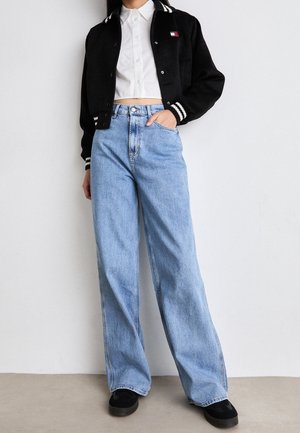 Personne portant un jean bleu clair à jambes larges, une chemise blanche courte boutonnée, une veste universitaire noire et des baskets noires, debout contre un mur blanc.