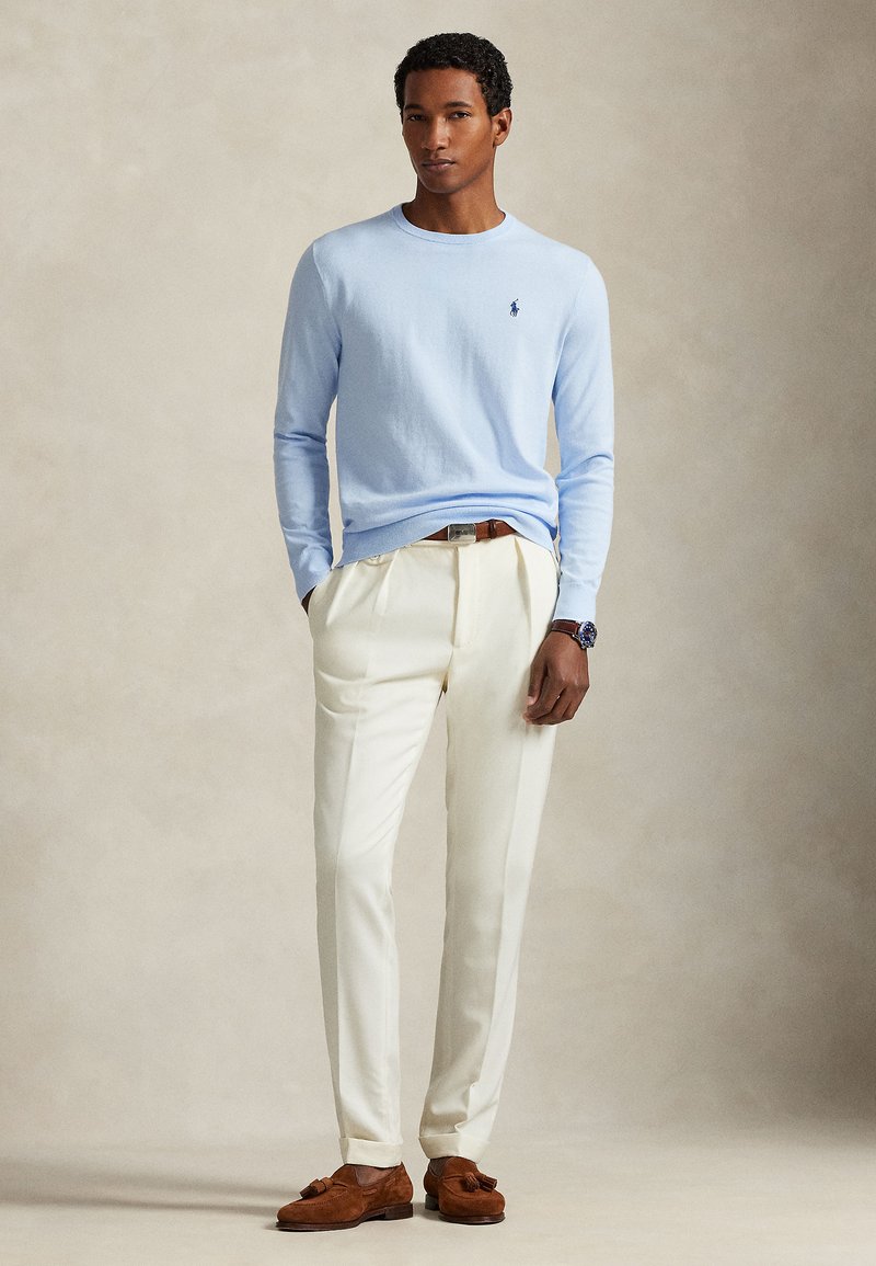 Maglione a maglia azzurro chiaro, pantaloni beige sartoriali e mocassini in suede marrone. Semplice scollo rotondo, con un piccolo logo sul petto.