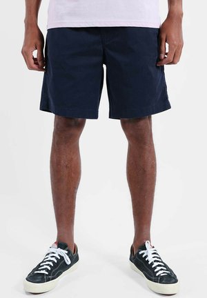 Une personne portant un short bleu marine, des baskets noires basses avec des lacets blancs et une chemise de couleur claire, debout sur un fond blanc.
