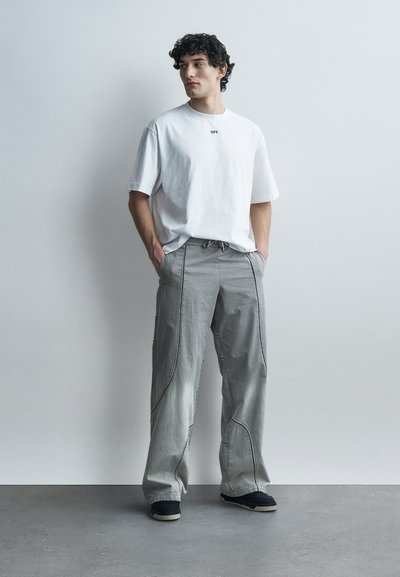 Νεαρός άνδρας με σγουρά μαλλιά που φοράει λευκό φαρδύ T-shirt και χαλαρό γκρι παντελόνι, στέκεται με τα χέρια στις τσέπες, κοιτάζοντας προς τα δεξιά.