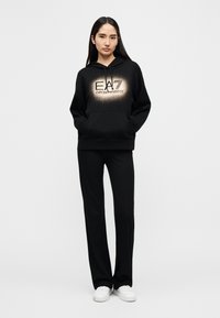 Μαύρο φούτερ με κουκούλα με μεγάλο χρυσό λογότυπο EAG Emporio Armani, σε συνδυασμό με μαύρο παντελόνι και λευκά αθλητικά παπούτσια. Καθημερινό στυλ με τσέπες στο φούτερ.