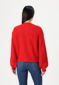 Femme aux longs cheveux noirs portant un pull rouge en maille avec des manches bouffantes et un jean bleu, vue de dos sur un fond blanc.