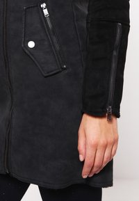 Giacca in finta suede nera con tasca con zip, dettagli con bottone a pressione e maniche testurizzate. Il design include cuciture e zip a vista.