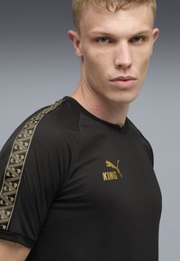 Sort atletisk t-shirt med struktureret stof, guldaccenter og et logo. Sidestriber viser et geometrisk guldmønster.