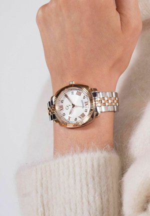 Orologio in metallo con cinturino in tonalità oro e argento, quadrante bianco, indicatori numerali romani e dettagli in cristallo; ghiera rotonda con bordo zigrinato.