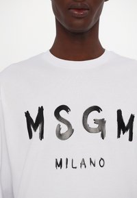 Gros plan d'une personne portant une chemise blanche avec des lettres noires style coups de pinceau "MSGM" et un plus petit "MILANO" en dessous.