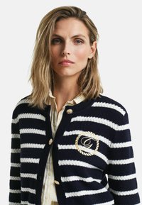 Marinblå och vit randig kofta med strukturerad stickning, guldknappar och en broderad logotyp i cirkulär design på vänster bröst.
