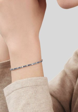 Wrist adorned with a delicate tennis bracelet featuring alternating blue and clear gemstones, worn with a beige knitted sleeve.
