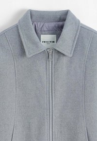 Veste gris mélange de laine avec une fermeture à glissière sur le devant, un col pointu et des coutures discrètes. Intérieur doublé d'un tissu violet clair. Étiquette de la marque visible.