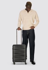 Hombre con suéter beige y jeans oscuros sosteniendo una maleta negra con ruedas y mango extendido contra un fondo gris.