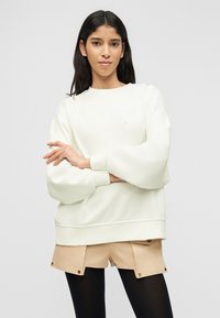 Νεαρή γυναίκα με ίσια σκούρα μαλλιά φοράει λευκό φαρδύ φούτερ, μπεζ σορτς με κουμπιά και μαύρες καλσόν, στέκεται με σταυρωμένα τα χέρια.