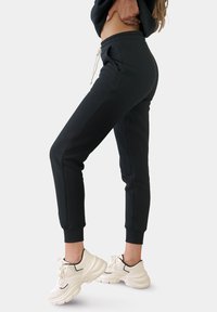 Zwarte trainingsbroek met een elastische tailleband en trekkoord, voorzien van zijzakken en taps toelopende benen, gecombineerd met chunky witte sneakers.