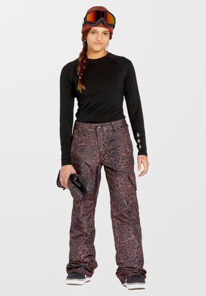 Completo da snowboard con una maglietta nera a maniche lunghe e pantaloni marroni con stampa leopardata, abbinato a un berretto rosso e occhiali da sci specchiati.