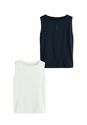 Frau mit lockigem Haar, die ein ärmelloses marineblaues Oberteil und schwarze Shorts trägt, vor einem schlichten weißen Hintergrund steht und nach unten schaut, dabei die Hand in der Nähe des Ohrs hält.