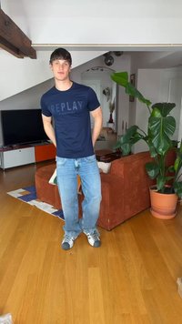 Νέος άνδρας με σκούρα μαλλιά που φοράει μια μπλε μπλούζα "REPLAY", ανοιχτόχρωμα τζιν και αθλητικά παπούτσια, στέκεται σε ένα σαλόνι με καφέ καναπέ και μεγάλο πράσινο φυτό.