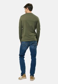 Chemise à manches longues vert olive, jeans en denim bleu et baskets beiges. La chemise a un col rond ; les jeans possèdent des poches arrière et un ajustement slim.
