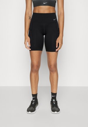 Atletische vrouw die zwarte hooggetailleerde Nike-shorts met zakken draagt, zwarte sokken, zwarte hardloopschoenen en een zwarte sportbeha, staand op een grijze vloer.