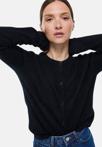 Cardigan noir en matière douce, avec un col rond, des manches longues et six boutons sur le devant. Associé à un jean bleu taille haute.