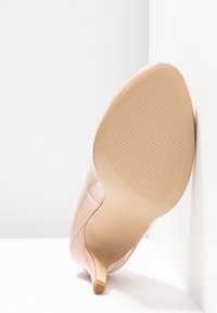 Escarpin en cuir verni beige avec un bout pointu et un talon aiguille élancé. La semelle présente une texture rainurée et un logo embossé.
