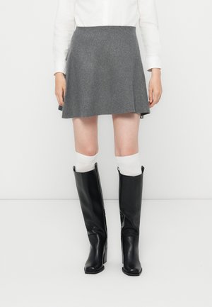 Personne portant une jupe grise, des chaussettes blanches montantes jusqu'aux genoux, des bottes noires montantes aux genoux et un haut blanc à manches longues, debout devant un fond uni.