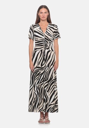Donna in piedi che indossa un vestito con stampa zebrata bianca e nera, maniche corte, scollo a V e dettaglio arricciato sul davanti, abbinato a sandali bianchi.