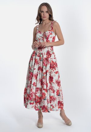 Donna in piedi che indossa un vestito floreale bianco senza maniche con fiori rossi e rosa, abbinato a scarpe beige con zeppa e cinturino alla caviglia, su sfondo semplice.