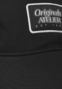 Fekete sapka strapabíró anyagból, fehér folttal, amelyen feketével hímzett "Originals ATELIER" és "EST. 199" felirat található. Finom textúra és varrás részletek.
